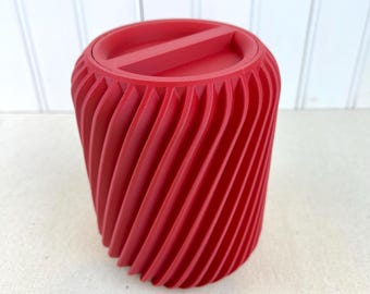 May include: A red cylindrical container with a textured, swirling design. The container has a lid with a small opening.