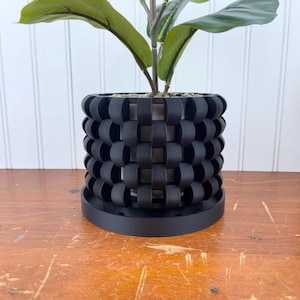 May include: A black plant pot with a woven design. The pot has a round base and is made of a sturdy material. A green plant is growing in the pot.