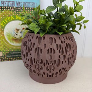 May include: A brown, spherical planter with a textured, porous design, filled with vibrant green artificial foliage. The planter sits on a matching base. A children's book is visible in the background.