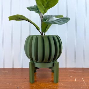 Op de afbeelding: Een groene keramische plantenpot met een geribbeld ontwerp staat op een bijpassende groene standaard. De pot is gevuld met een groene, bladerrijke plant.