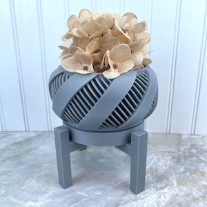 Puede incluir: Un macetero de cerámica gris con un diseño único y retorcido, con un soporte de tres patas. El macetero está lleno de flores secas beige.