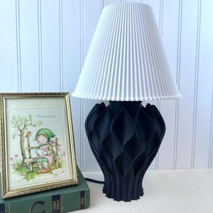 May include: A black table lamp with a white pleated lampshade. The lamp base is made of a geometric design with a repeating pattern of leaves. The lamp is sitting on a book with a gold-colored frame on top of it.