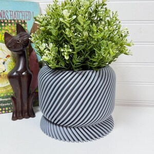 May include: A grey and white striped planter with a green plant. The planter has a unique design with diagonal lines and a matching base. A dark brown cat figurine and a vintage book are in the background.