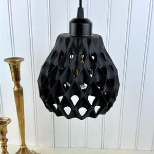 Puede incluir: Una lámpara colgante negra con una pantalla geométrica con forma de diamante. La pantalla tiene espacios abiertos que permiten que la luz brille. La lámpara está suspendida de un cable negro, contra una pared blanca con paneles verticales.