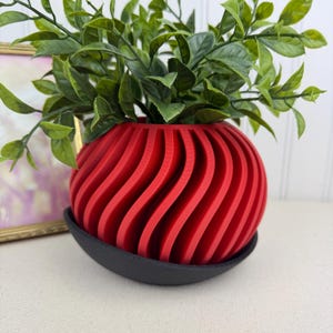 May include: A red, spherical planter with a unique, wavy design, holding green leafy plants. The planter sits on a black saucer. The design features vertical, curved lines, creating a modern aesthetic. A decorative item for home decor.