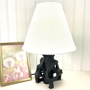 Può includere: Una lampada da tavolo nera con paralume bianco plissettato. La lampada ha una base astratta e scultorea. Un'immagine incorniciata con una cornice dorata è a sinistra della lampada.
