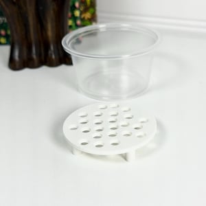 Puede incluir: Un recipiente de plástico transparente con tapa se encuentra encima de una pequeña plataforma circular blanca con numerosos agujeros espaciados uniformemente. La plataforma tiene tres patas cortas. Los artículos están sobre una superficie blanca.