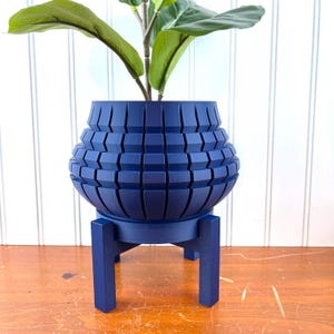 Op de afbeelding: Een marineblauwe geometrische plantenbak met bijpassende standaard, met een groene plant. De plantenbak heeft een uniek, gestructureerd ontwerp. De standaard heeft een ronde basis en vier poten. De plant heeft grote, groene bladeren.