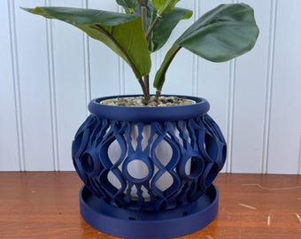 May include: A navy blue decorative plant pot with a unique openwork design. The pot holds a green plant with large leaves. The pot sits on a matching navy blue saucer. The pot's design features a series of interconnected oval shapes.