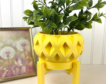 May include: A yellow plant pot with a diamond pattern, containing a green plant. The pot is on a matching yellow stand. A framed picture with a gold frame is in the background. The pot's diameter is approximately 20 cm.