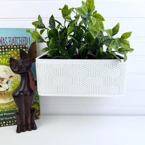 Puede incluir: Una maceta de pared rectangular blanca con un patrón geométrico en relieve de formas en Y entrelazadas, que contiene una planta verde. Al lado hay una figura de gato de madera marrón oscuro junto a un libro titulado "Was Hatched!" de Cyndy Szekeres.