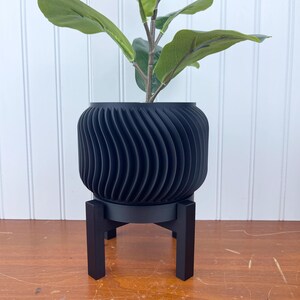 Puede incluir: Una maceta negra con un diseño ondulado y acanalado se asienta sobre un soporte de madera negro. La maceta está llena de una planta verde con hojas grandes.