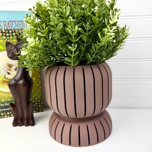May include: A mauve-colored planter with a rounded, ribbed design featuring vertical cutouts along the body and base. The planter holds a lush, artificial green plant with small, rounded leaves that extend outward from the top opening.