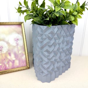 May include: A light gray geometric planter with a textured surface, filled with green leafy plants. The planter has a unique angular design. A framed picture with a gold frame is to the left of the planter.
