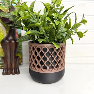 May include: A cylindrical plant pot featuring a matte black base and a contrasting dusty rose upper section with an intricate, repeating lattice cutout pattern. A vibrant green leafy plant is potted inside the container, which sits next to a dark brown cat figurine.