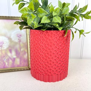 Puede incluir: Una maceta cilíndrica roja vibrante con un patrón geométrico, que contiene una planta verde. La maceta tiene una superficie texturizada y una base decorativa. Una imagen enmarcada con un marco dorado está en el fondo.