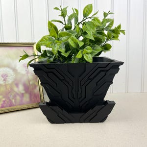 May include: A black square planter with a geometric design, holding a vibrant green plant. The planter has a matching saucer. The design features angular lines and shapes, creating a modern aesthetic. The plant adds a natural contrast to the dark planter.