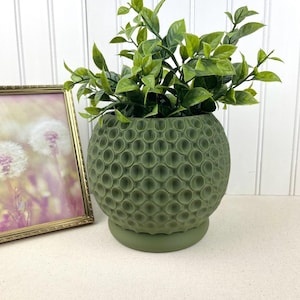 Puede incluir: Una maceta esférica verde texturizada con una base de platillo a juego, llena de vibrantes plantas artificiales de hojas verdes. Una imagen enmarcada con un borde dorado es visible en el fondo, agregando un toque decorativo.
