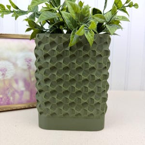 May include: An olive green geometric patterned planter filled with green leafy plants. The planter has a textured surface with a repeating honeycomb design. The base of the planter is a solid olive green.