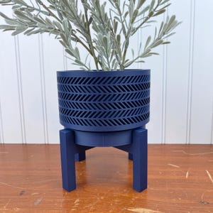 May include: A navy blue planter with a geometric pattern sits on a matching navy blue stand with three legs. The planter is filled with a green leafy plant.