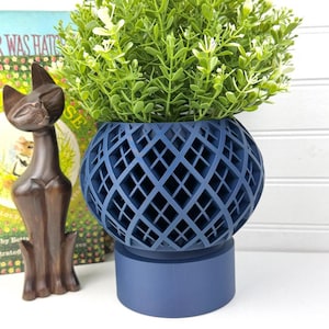 May include: A dark blue spherical planter with a geometric lattice pattern and a solid cylindrical base. The planter holds a lush green artificial plant with small white flowers. A dark brown wooden cat figurine stands to the left of the planter.