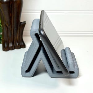 May include: A gray phone stand holds a black smartphone. The stand has a zigzag design with a series of small, rounded supports to hold the phone. The stand is made of a 3D-printed material.