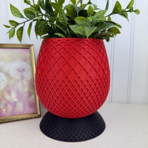 May include: A red, diamond-patterned planter with a black base, holding green leafy plants. The planter has a textured surface and a rounded shape. A framed picture is visible in the background.