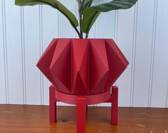 May include: A red geometric planter with a stand. The planter has a starburst design and is made of ceramic. The stand is also red and has a simple design.