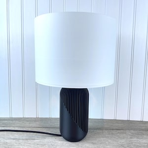 Op de afbeelding: Een moderne tafellamp met een witte cilindrische lampenkap. De voet is zwart met een diagonaal ontwerp en verticale lijnen. De lamp is aangesloten op een zwart netsnoer.