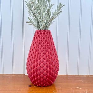 May include: A red ceramic vase with a textured, diamond-shaped pattern. The vase is shaped like a teardrop and is filled with a sprig of dried greenery.