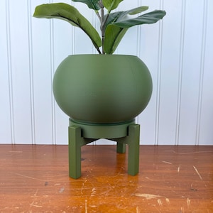 May include: A green, spherical planter with a plant, set on a matching green stand. The planter has a smooth surface and a round shape. The stand has a circular base and four square legs. The plant has large, green leaves.