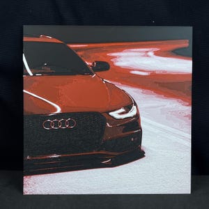 May include: A square print featuring a red Audi car. The car's front is prominently displayed, with the Audi logo visible on the black grill. The background shows a blurred red and white design, suggesting speed and motion.