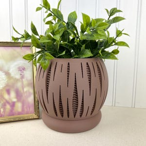 May include: A decorative, round, mauve-colored planter with a unique cut-out design, holding vibrant green leafy plants. The planter sits on a matching base. A framed picture with a gold frame is visible in the background.
