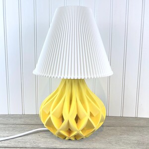 May include: A yellow geometric table lamp with a white pleated lampshade. The lamp has a unique, modern design with a geometric pattern.