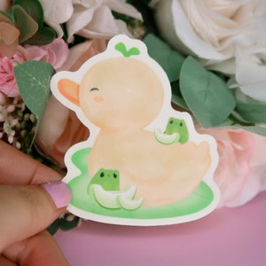 Puede incluir: Un pato amarillo con una hoja verde en la cabeza está rodeado de dos ranas verdes en un nenúfar verde. El pato tiene una expresión feliz y las ranas tienen ojos negros.