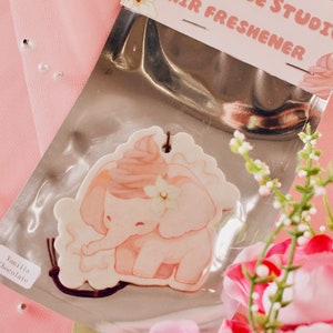 Könnte beinhalten: Ein rosa-weißer Elefanten-Lufterfrischer mit Vanille-Schokoladen-Duft. Der Elefant hält eine Blume und hat ein rosa-weißes Frosting-artiges Design auf seinem Kopf. Der Lufterfrischer ist in einer durchsichtigen Plastiktüte mit einer braunen Schnur verpackt.