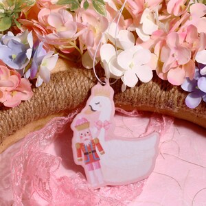 May include: A pink and white porcelain figurine of a nutcracker and a swan. The nutcracker is wearing a red and gold uniform and holding a sword. The swan is wearing a crown and has its wings spread out. The figurine is surrounded by pink and white flowers.