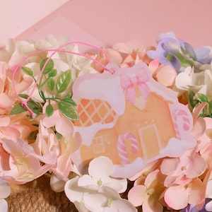 Op de afbeelding: Een roze en witte gingerbread house-hanger met een roze strik en snoepstokaccenten. De hanger is omringd door een boeket pastelroze, witte en paarse bloemen.