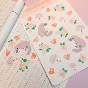 May include: A sheet of stickers featuring a cartoon platypus, flowers, hearts, and umbrellas. The stickers are on a white background and have a pink and peach color scheme. The text "Foxy Rose Studio I Love U" is at the top of the sheet.