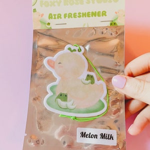 Puede incluir: Un ambientador blanco y verde con un pato y ranas de dibujos animados. El ambientador está empaquetado en una bolsa de plástico transparente con una cuerda verde. El texto "Foxy Rose Studio" y "Air Freshener" está impreso en el embalaje. El texto "Melon Milk" está impreso en una etiqueta blanca.