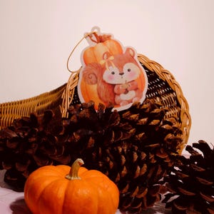 May include: A brown wicker cornucopia filled with pine cones and a small orange pumpkin. A paper cutout of a squirrel holding a pumpkin is attached to the cornucopia.