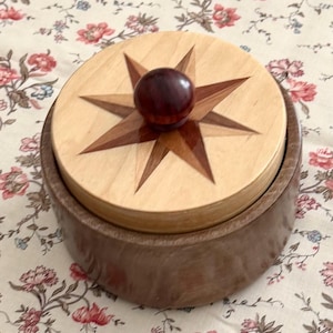 May include: A round wooden container with a dark brown base and a light tan lid. The lid features an inlaid dark wood star design radiating from the center, topped with a small, spherical, dark reddish-brown wooden knob handle.