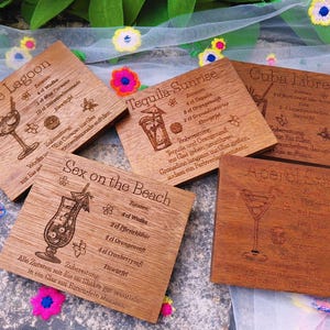 May include: Wooden coasters with engraved cocktail recipes, including "Blue Lagoon," "Tequila Sunrise," and "Sex on the Beach." Each coaster features a drink illustration and ingredient list. The coasters are square and made of light brown wood.