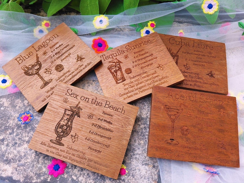 May include: Wooden coasters with engraved cocktail recipes, including "Blue Lagoon," "Tequila Sunrise," and "Sex on the Beach." Each coaster features a drink illustration and ingredient list. The coasters are square and made of light brown wood.