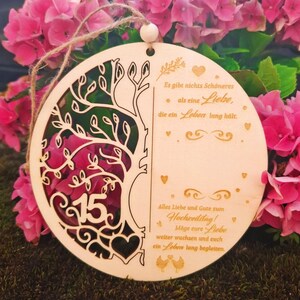 K&ouml;nnte beinhalten: Runder Holzschmuck mit einem Baumdesign und der Zahl 15. Der Schmuck hat ein ausgeschnittenes Design und enth&auml;lt deutschen Text. Er wird mit einer Schnur aufgeh&auml;ngt und ist von rosa Blumen umgeben.