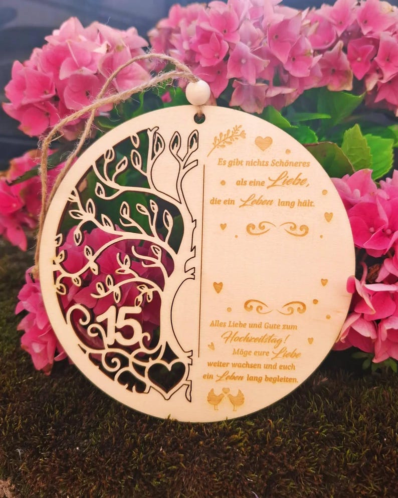 K&ouml;nnte beinhalten: Runder Holzschmuck mit einem Baumdesign und der Zahl 15. Der Schmuck hat ein ausgeschnittenes Design und enth&auml;lt deutschen Text. Er wird mit einer Schnur aufgeh&auml;ngt und ist von rosa Blumen umgeben.