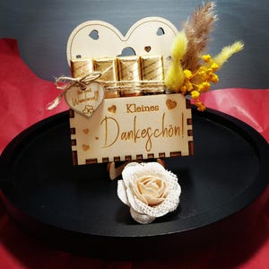 May include: A wooden gift box with the words "Kleines Dankeschön" and a heart-shaped cutout. The box holds small, gold-wrapped chocolates and is decorated with dried flowers and a fabric rose. The box sits on a black tray.