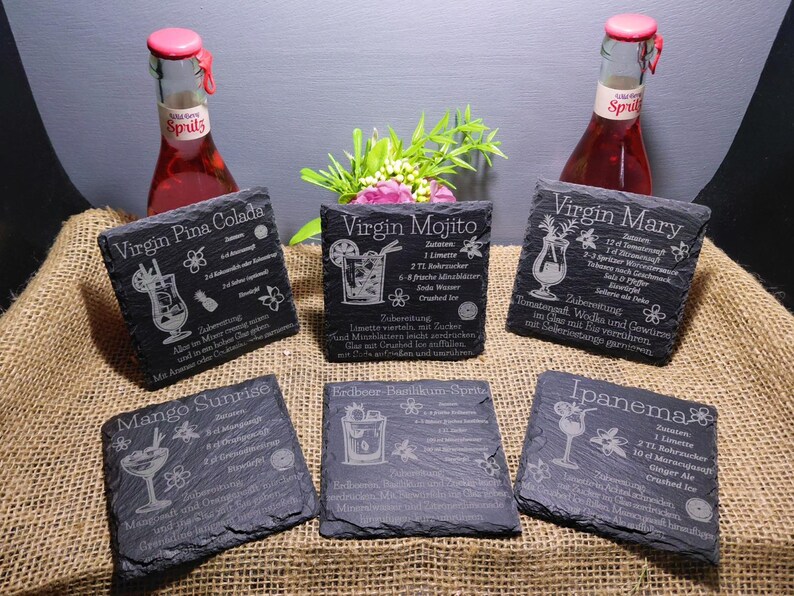 May include: A collection of six dark gray slate coasters with engraved cocktail recipes, including "Virgin Pina Colada," "Virgin Mojito," and "Virgin Mary." Two bottles of red spritz are in the background.