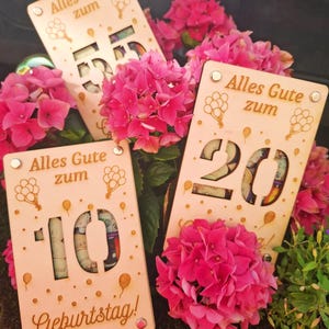 May include: Wooden birthday gift cards with cut-out numbers 10, 20, and 55, filled with rolled-up bills. The cards have the German text "Alles Gute zum Geburtstag!" and balloon graphics. Bright pink hydrangea flowers surround the cards.