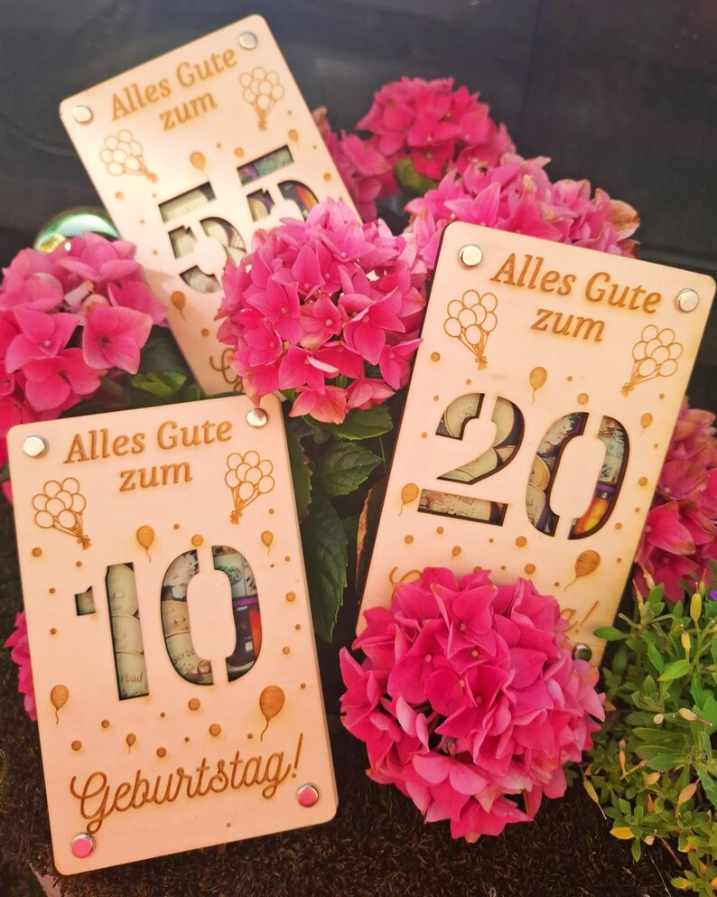 May include: Wooden birthday gift cards with cut-out numbers 10, 20, and 55, filled with rolled-up bills. The cards have the German text "Alles Gute zum Geburtstag!" and balloon graphics. Bright pink hydrangea flowers surround the cards.